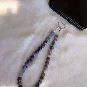 Peut inclure: Une dragonne de téléphone composée d'un cordon de perles rondes et polies dans des tons variés de bleu, gris et blanc. Les perles sont enfilées et fixées à un fermoir mousqueton en métal argenté et à une boucle de connexion en plastique transparent.