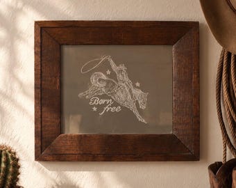 May include: A dark brown wooden frame containing a glass panel with a white etched illustration of a cowboy riding a bucking horse while swinging a lasso. The text Born free is etched below the horse, accompanied by two small star shapes.