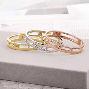 Könnte beinhalten: Drei aufklappbare Armreifen in Gold-, Silber- und Roségoldtönen. Jeder Armreif hat ein Doppelband-Design mit einer mittleren Reihe aus kleinen, klaren, rund geschliffenen Steinen und drei größeren, beweglichen, gefassten, runden Steinen.