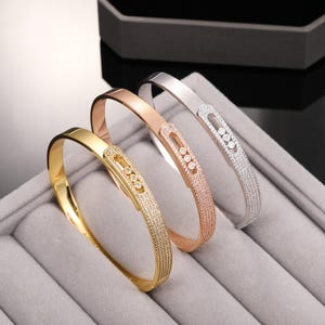 Könnte beinhalten: Drei aufklappbare Armreifen in Gold-, Roségold- und Silbertönen. Jeder Armreif verfügt über einen rechteckigen Mittelteil, der mit kleinen klaren Steinen besetzt ist, sowie drei runde Steine in einer vertikalen Fassung mit Pavé-Besatz.