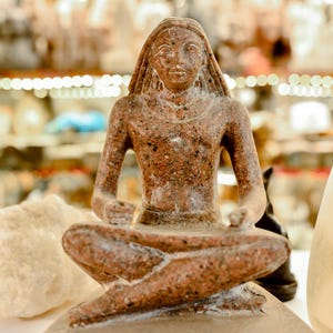 May include: A speckled brown granite sculpture of a seated figure in a cross-legged position. The figure has a smooth, polished surface with visible mineral flecks, a defined facial structure, and hair that falls to the shoulders, resting on a base.
