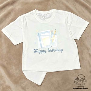 May include: A white short-sleeved t-shirt featuring a graphic of a clear pencil case containing a notebook, a yellow sticky note, and two pencils. Below the graphic, the text "Happy learning" is printed in a blue cursive script font.