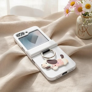 May include: A white Samsung Galaxy Z Flip6 phone case featuring a silver metal ring holder and a flat, cartoon-style dog figure with brown ears and a pink polka-dot pattern on its back. The case has cutouts for the camera lenses and the screen.