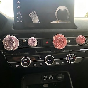 May include: Four flower-shaped car air vent clips attached to the dashboard vents. Two clips are light purple peonies and two are pink peonies. The car dashboard features a digital screen displaying an astronaut waving and climate control dials.