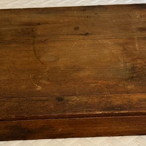 May include: A rectangular wooden box with a dark brown finish and a flat lid. The surface of the box shows visible wood grain, natural knots, and various surface scratches, indicating a weathered and aged appearance throughout the exterior.