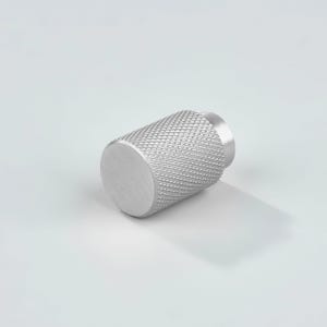 May include: A cylindrical metal knob with a brushed silver finish and a textured diamond knurled pattern along the side. The front face is smooth and flat, while the back features a smaller circular base for mounting purposes.