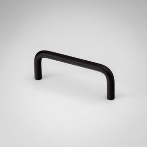 Puede incluir: Un tirador de mueble en color negro mate con un diseño sencillo en forma de U redondeada. El herraje presenta un acabado liso y uniforme con dos puntos de montaje verticales en cada extremo, creando una estética limpia y funcional.