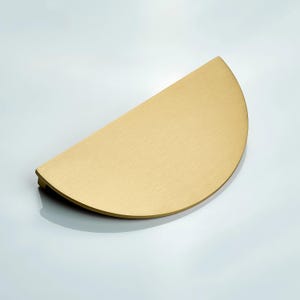 May include: A semi-circular cabinet pull with a brushed gold finish. The hardware features a flat, curved edge and a straight mounting side, showcasing a smooth, metallic texture that reflects light across its surface in a simple, functional design.