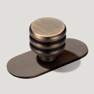 May include: A round cabinet knob with a brushed bronze finish featuring three horizontal ridges, mounted on a smooth, oval-shaped backplate. The metal hardware has a dark, metallic tone with visible horizontal graining across the surface.