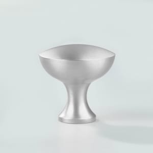 Op de afbeelding: Een meubelknop met een geborstelde zilveren afwerking, voorzien van een breed, afgerond bovenoppervlak dat taps toeloopt in een smalle, gebogen hals en een ronde voet. Het metalen beslag heeft een gladde, matte textuur en grijze kleur.