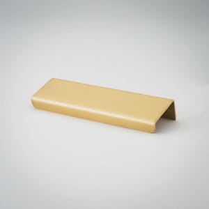 May include: A gold-colored metal cabinet pull with a smooth, matte finish. The handle features a rectangular, elongated shape with a curved front edge that folds downward, creating a simple, functional design for drawers or cabinet doors.