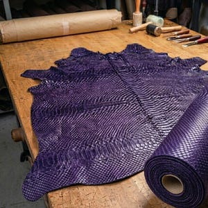 May include: A large, deep purple piece of textured snakeskin leather with a distinct scale pattern, partially laid flat and partially rolled into a cylinder. The material displays a consistent, vibrant violet hue across its entire surface.