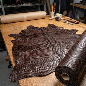 Peut inclure: Une peau de cuir de serpent brun foncé avec un motif d'écailles distinct posée à plat sur un établi en bois, accompagnée d'une section roulée du même matériau texturé, mettant en valeur la texture reptilienne naturelle et les tons bruns.