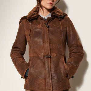 May include: A brown suede jacket with a dark brown faux fur collar and hood. The jacket features a front closure with two toggle buttons, long sleeves, and two side pockets. The material has a distressed texture and a visible seam across the waist.