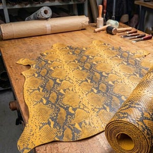 Puede incluir: Una pieza grande y plana de cuero amarillo y gris oscuro con estampado de piel de serpiente extendida sobre un banco de trabajo de madera, acompa&ntilde;ada de una secci&oacute;n enrollada del mismo material, con un dise&ntilde;o de escamas texturizado.