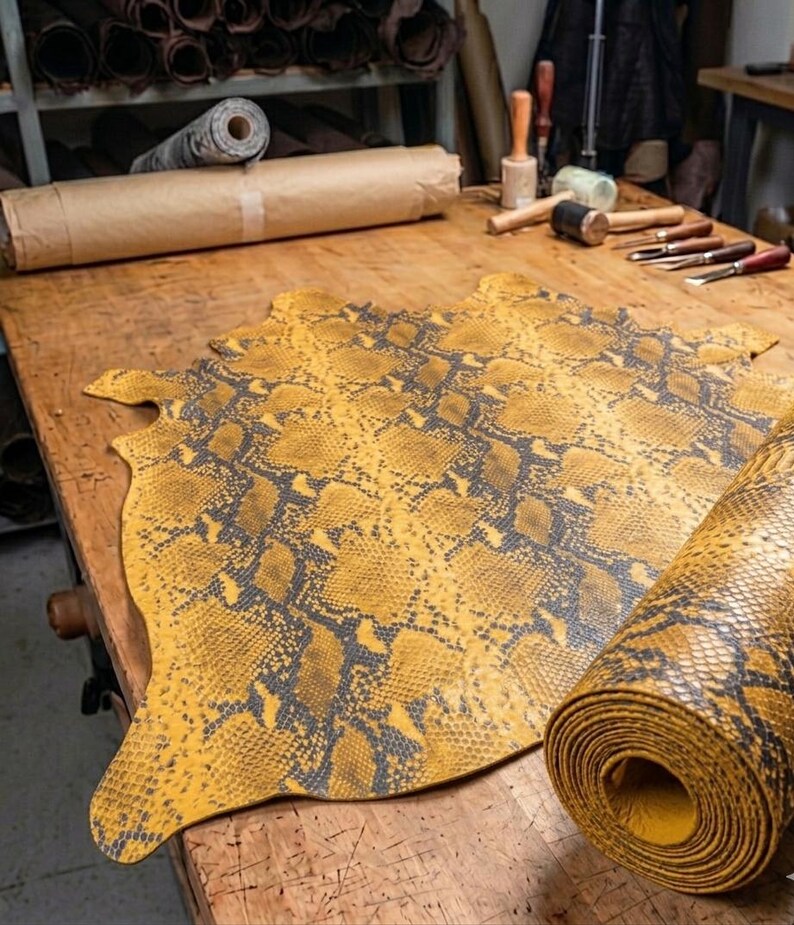 Puede incluir: Una pieza grande y plana de cuero amarillo y gris oscuro con estampado de piel de serpiente extendida sobre un banco de trabajo de madera, acompa&ntilde;ada de una secci&oacute;n enrollada del mismo material, con un dise&ntilde;o de escamas texturizado.