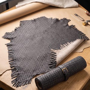 May include: A dark charcoal grey woven leather hide with a fringed edge, accompanied by a matching rolled leather piece secured with a black strap and a brass buckle. The texture features a consistent grid pattern throughout the material.
