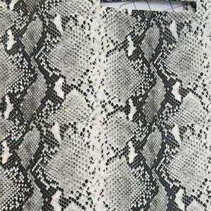 Puede incluir: Una l&aacute;mina de material con un patr&oacute;n de piel de serpiente en gris, negro y blanco. La superficie muestra una textura de escamas repetitiva con un dise&ntilde;o en zigzag. Dos recortes rectangulares horizontales son visibles en el borde superior.