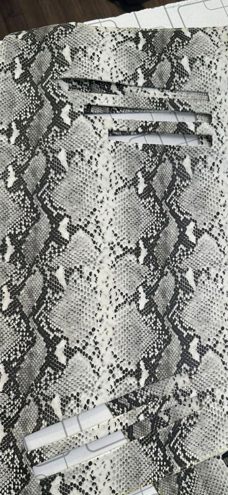 Puede incluir: Una l&aacute;mina de material con un patr&oacute;n de piel de serpiente en gris, negro y blanco. La superficie muestra una textura de escamas repetitiva con un dise&ntilde;o en zigzag. Dos recortes rectangulares horizontales son visibles en el borde superior.