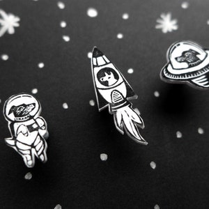 May include: Three black and white enamel pins featuring a dog astronaut, a dog in a rocket ship, and a dog in a flying saucer. The pins are set against a black background with white dots.