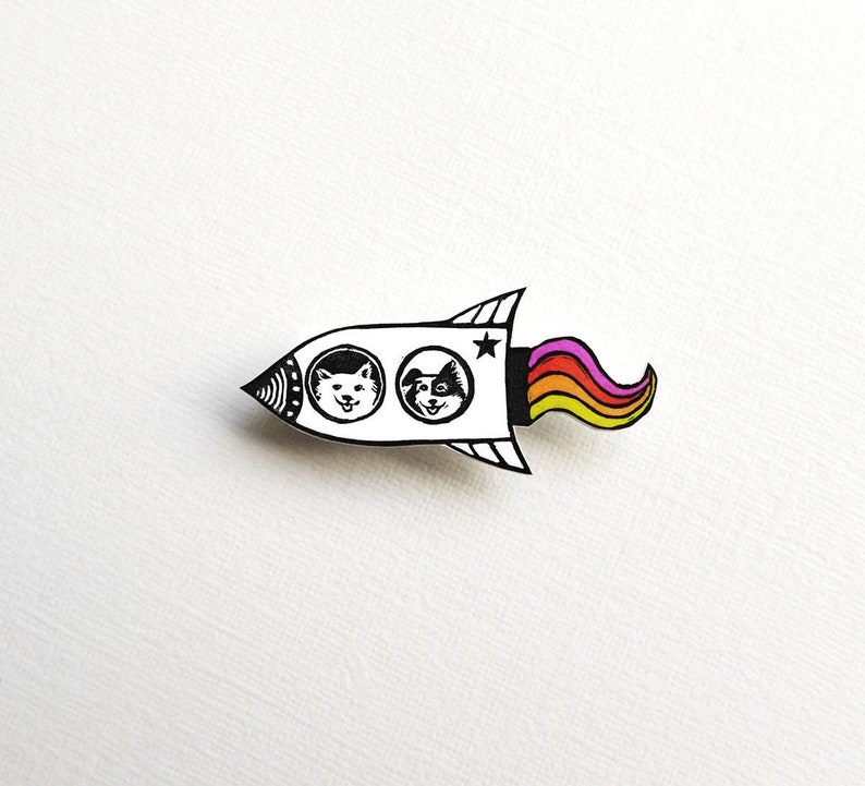 May include: A white rocket ship with two black and white dog faces in the windows. The rocket has a rainbow-colored tail flame.