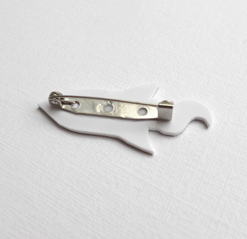 May include: White rocket ship shaped pin with a silver metal pin back.