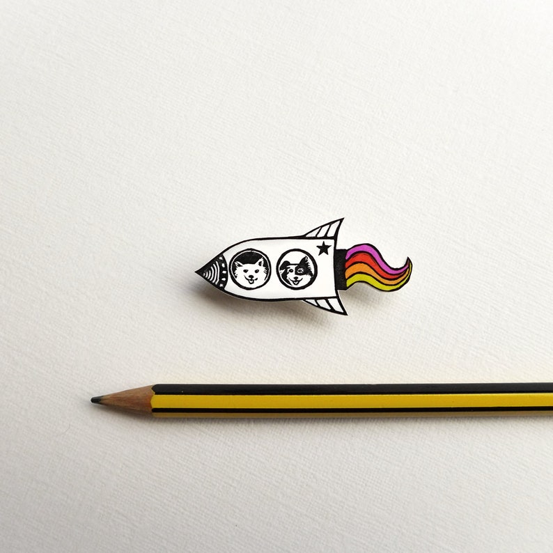 May include: A white rocket with black outlines and a rainbow tail. Two black and white animal faces are visible in the windows of the rocket.