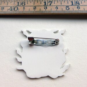 May include: White ceramic pin with a detailed design of a creature with a pin back.