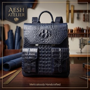 Può includere: Uno zaino blu scuro con un motivo testurizzato in pelle di coccodrillo, dotato di manico superiore, due tasche frontali con chiusura a patta e spallacci. La borsa riporta il testo AESH ATELIER e Meticulously Handcrafted.
