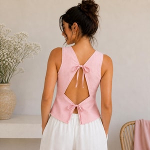 Könnte beinhalten: Ein ärmelloses, hellrosa Oberteil mit offenem Rückendesign und zwei dünnen Stoffbändern am Hals und an der Taille. Das Kleidungsstück wird mit einer weißen Hose kombiniert und zeigt eine klare Ästhetik mit sichtbarer Stoffstruktur und Passform.
