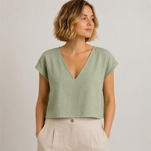 May include: A sage green short-sleeved top with a deep V-neckline and a boxy, cropped silhouette, paired with high-waisted beige trousers featuring a single visible button closure at the centre front waistband and subtle pleat detailing.