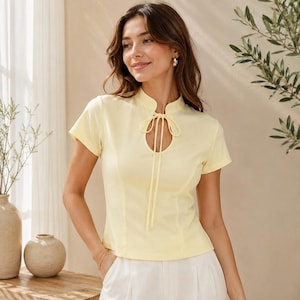 May include: A pale yellow short-sleeved top featuring a mandarin collar, a teardrop-shaped cutout at the neckline, and two long, thin fabric ties hanging from the collar. The garment has a fitted silhouette with vertical seam detailing.