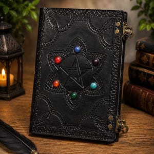 May include: A black leather journal featuring an embossed pentagram design surrounded by six colorful inlaid stones. The cover includes intricate border patterns and two brass-colored metal latches on the side to secure the book closed.