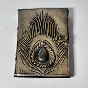 May include: A rectangular journal with a distressed metallic bronze leather cover featuring an embossed peacock feather design. A teardrop-shaped iridescent stone is set in the center, surrounded by a braided border, and secured with a brass latch.