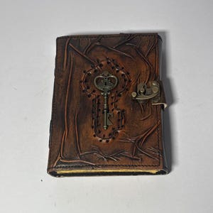 May include: A rectangular brown leather journal featuring a textured, tree-like embossed pattern on the cover. A metal key emblem is centered within a stitched outline, and a brass-colored metal latch secures the side of the book.