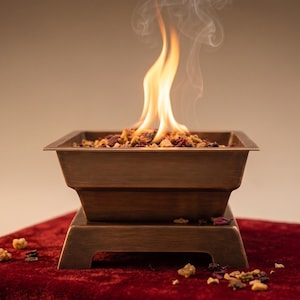 May include: A square, bronze-coloured metal fire pit bowl with a stepped base design. The bowl is filled with a mixture of dried botanical elements and small wood pieces, with bright orange flames and wisps of smoke rising from the centre.