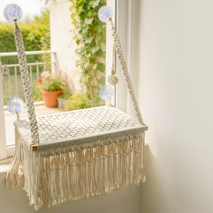 Pode incluir: Um poleiro para gatos em macramé cor de creme com uma plataforma retangular, superfície superior tecida e franjas longas. O poleiro é suspenso por duas cordas de macramé trançadas fixadas ao vidro da janela com quatro ventosas.