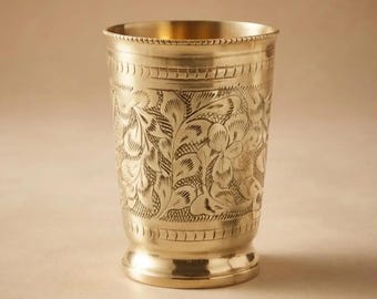 Può includere: Un bicchiere cilindrico in ottone caratterizzato da un intricato motivo floreale inciso attorno al centro. Il recipiente presenta un bordo liscio e lucido e una base svasata, con fasce decorative orizzontali che delimitano il disegno botanico.