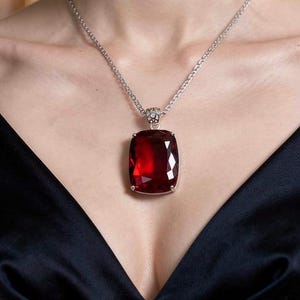 May include: A silver chain necklace featuring a large, rectangular, faceted deep red gemstone pendant. The pendant is set in a silver frame with a decorative bail, resting against a dark navy blue satin garment with a V-neckline and dark embellishments.