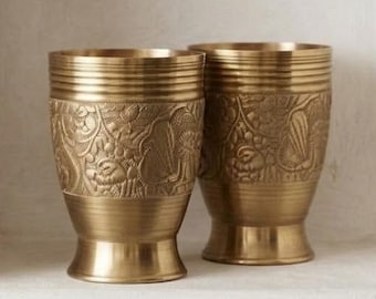 Può includere: Due tazze in ottone abbinate caratterizzate da un motivo floreale in rilievo sulla parte inferiore e dettagli a coste orizzontali sul bordo superiore. Ogni tazza presenta una base svasata e una finitura metallica dorata.
