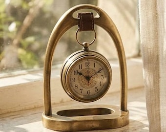 Può includere: Un orologio da tavolo in metallo color ottone sospeso a una cornice ad arco tramite un piccolo cinturino in pelle marrone. Il quadrante rotondo presenta numeri arabi neri e intricati motivi decorativi al centro, fissato su una robusta base a U.