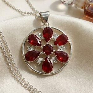 May include: A silver-toned necklace featuring a circular pendant with a central round red gemstone surrounded by six pear-shaped red gemstones arranged in a floral pattern, accented with small silver leaf-like details between the stones.