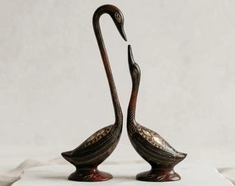Può includere: Due statuette di cigno in metallo marrone scuro rivolte l'una verso l'altra. Ogni cigno presenta intricati motivi argentati incisi lungo le ali e il corpo, con colli lunghi e sottili e una finitura lucida e liscia sulla base e sulla testa.