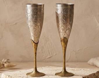 Può includere: Due flûte da champagne alte con coppe color argento caratterizzate da intricati motivi floreali incisi e steli con basi circolari in ottone a contrasto. I recipienti in metallo presentano una forma slanciata e una finitura lucida.