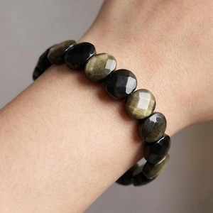 Könnte beinhalten: Ein elastisches Armband bestehend aus abwechselnd facettierten, ovalen Perlen in solidem Schwarz und schimmerndem, metallisch goldenem Obsidian. Die Perlen sind in einem durchgehenden kreisförmigen Muster angeordnet und haben eine polierte Oberfläche.