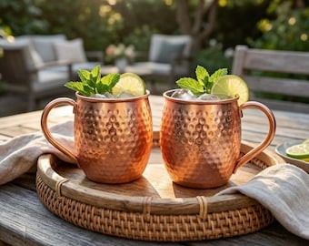 Può includere: Due tazze in rame martellato con manici curvi, ciascuna riempita con ghiaccio, una fetta di lime e foglie di menta fresca. Le tazze sono posizionate su un vassoio rotondo in legno intrecciato con un tovagliolo di stoffa chiara.
