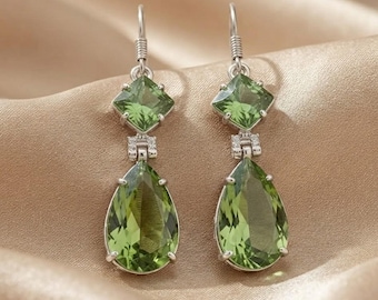 Può includere: Un paio di orecchini pendenti in argento con una gemma verde quadrata sfaccettata in alto e una gemma verde sfaccettata a forma di pera più grande in basso, collegate da un piccolo elemento in argento con minuscole pietre chiare incastonate.