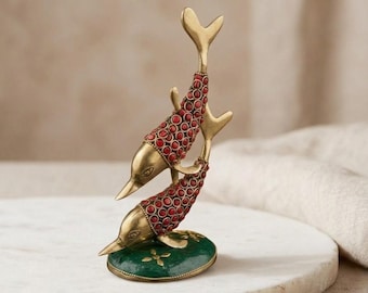 Può includere: Una scultura in ottone raffigurante due delfini impilati verticalmente. I corpi dei delfini sono intarsiati con piccole pietre rotonde rosse. La scultura è montata su una base circolare verde scuro con un piccolo dettaglio floreale dorato.