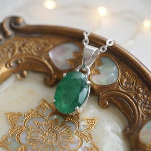 Elegant 10–15Ct Emerald Pendant Necklace featuring a natural green emerald in a classic oval cut. The gemstone displays beautiful natural variations and subtle light reflections that emphasize its authentic character. Set against a delicate silver-tone chain, this minimalist May birthstone necklace makes a versatile accessory for casual and formal fashion and a thoughtful gift for women, gemstone collectors, May birthdays, and emerald jewelry enthusiasts.