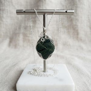 Natural certified green jade pendant displayed as a handcrafted wire-wrapped necklace with a polished crystal body, custom silver-tone wire framing, and neat loop details. The dark green stone provides a focal point while the artistic wire wrapping adds a refined decorative character. This unique necklace is suitable for crystal lovers, gemstone collectors, spiritual jewelry styling, statement accessories, special occasions, and thoughtful holiday or birthday gifts with an organic feel.
